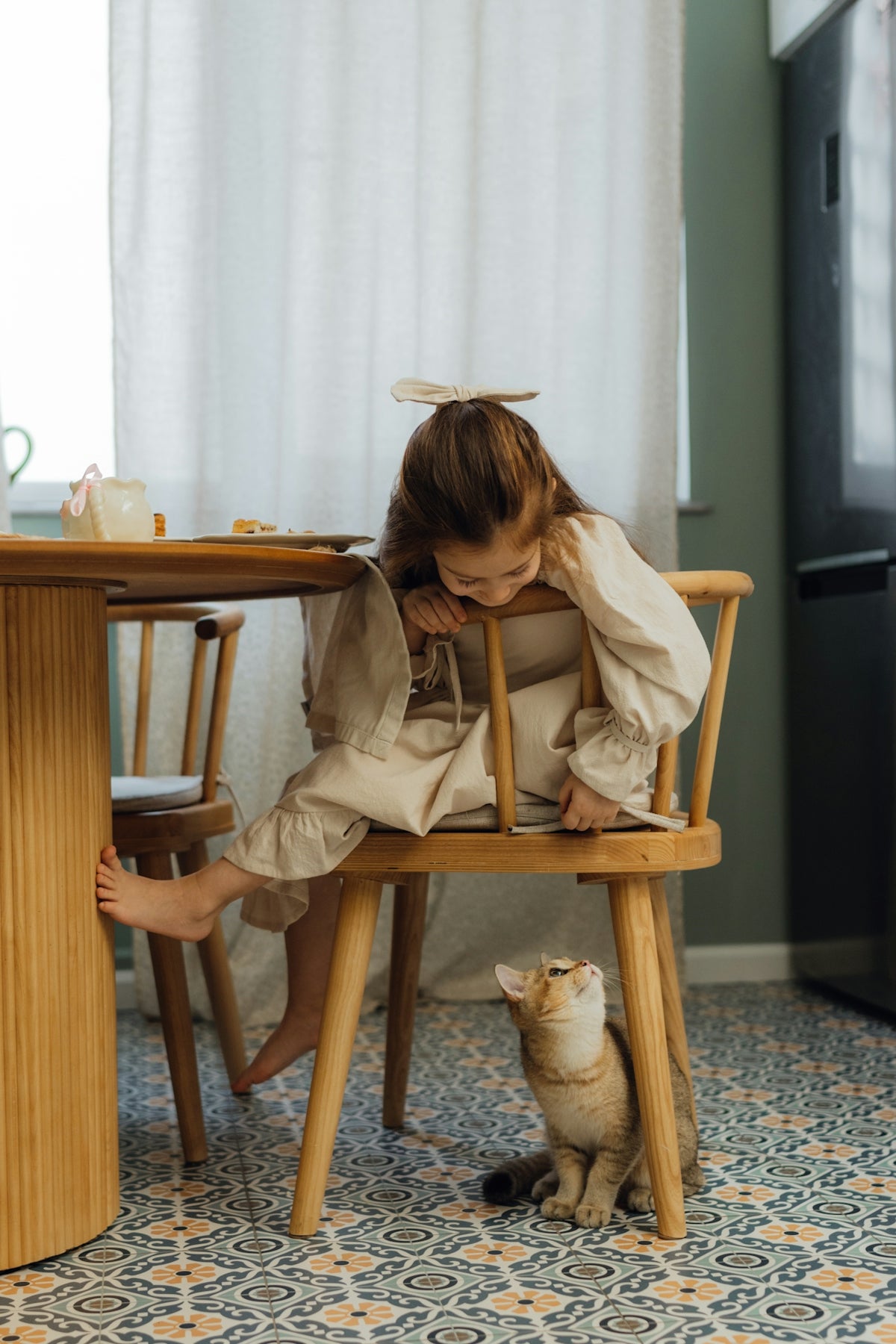 A little girl sitting in a wooden chair next to a cat